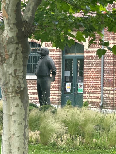 A statue of a man with a large nose and a prominent belly stands to the right of a large tree trunk, in front of a brick building with green doors and windows. The statue is dark in color, with the man wearing a suit and holding his right hand behind his back. The tree trunk in the foreground is large and textured, with a mottled appearance of grey, green, and brown. The building behind the statue is constructed of red brick and features a large, arched window with bars, and a double green door. The area in front of the building is landscaped with tall, wispy grasses and some smaller flowering plants. The statue is likely located in the city of Hoeilaart, Belgium, as it is identified as the "Statue of Papa Smet" in the city.