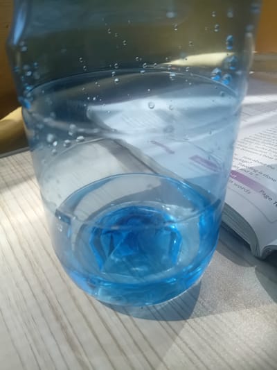 This image shows a close-up of a clear plastic bottle partially filled with blue liquid. The bottle is placed on a wooden surface with visible wood grain. A book with text and page numbers is visible to the right of the bottle, partially obscured. The lighting suggests an indoor environment with sunlight casting shadows. There are no people or animals present in the image. The location in Vidin, Bulgaria cannot be determined from the visual information provided.
