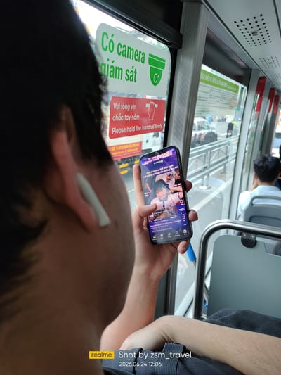 A person holds a smartphone while sitting on a bus. The phone displays a video or stream with Vietnamese text. Behind the person, a green and white sign is visible with Vietnamese text that translates to "Caution, camera surveillance." Below this, a red sign reads "Please hold the handrail firmly." The bus interior shows gray seats and windows looking out onto a street with vehicles. The lower right corner of the image includes a "realme" logo and text indicating the photo was taken by "zsm_travel" on "2026.06.24 12:06." The environment suggests this is within the city of Thuận An, Vietnam, a common location for public bus transportation.