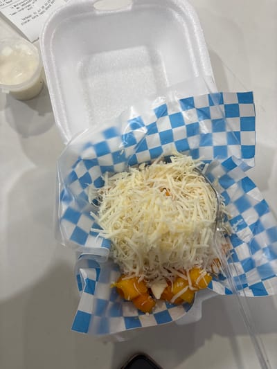 A top-down, close-up shot captures a serving of food presented in a white styrofoam container lined with blue and white checkered paper. The food itself is topped with a generous amount of shredded cheese, beneath which are visible pieces of what appear to be sliced mangoes. A clear plastic fork rests on the right side of the container, partially submerged in the food. To the left of the container, a small, clear plastic cup filled with a white, creamy substance is visible, along with a receipt. The background is a plain, white surface, likely a table. The receipt indicates a transaction on "01 Jul 26 20:23" and mentions "Temen nongkrong" and "ADMIN TANSU GRAHA RAYA sini," which suggests the location is likely in the Graha Raya area of South Tangerang, Indonesia, a popular neighborhood known for its restaurants and cafes.