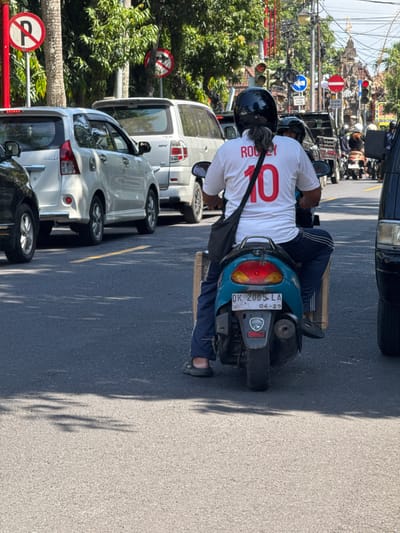 The image captures a street scene in Gianyar, Indonesia, during daylight hours, likely mid-morning or afternoon given the harsh shadows. The foreground is dominated by a person riding a teal-colored scooter. The rider, wearing a black helmet and a white t-shirt with red lettering and the number "10" on the back, is seen from behind. They are also wearing dark blue pants with white stripes. A black shoulder bag is slung across their chest. The scooter's license plate is visible and reads "DK 2005 LA" with a date "04-29" below it. Cardboard boxes are strapped to the rear of the scooter.

The scooter is in traffic, surrounded by other vehicles. To the left, a white car is partially visible. Behind the scooter, other motorcycles and cars are present, suggesting a busy urban environment. Traffic lights and street signs are visible in the background, indicating an intersection or main road. Trees line the street, their foliage creating dappled shadows on the asphalt. The overall impression is of a typical traffic scene in a bustling Indonesian city.