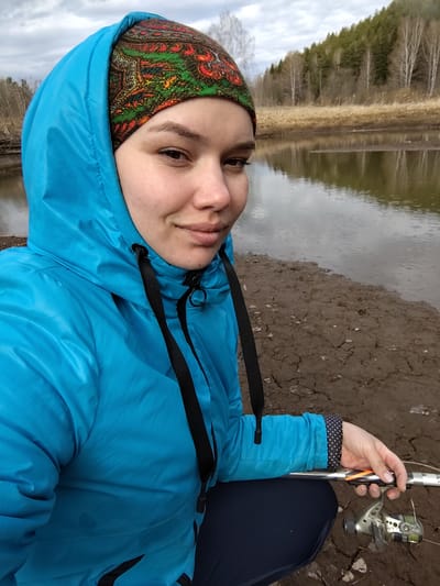 A young adult woman takes a selfie outdoors by a body of water, likely a river or large creek, in what appears to be Kresty, Russia. She is positioned close to the muddy bank, which shows signs of cracking, suggesting it might have been wet and then dried.
The woman has a light complexion and dark eyebrows, looking directly at the camera with a slight, calm smile. She wears a bright, almost neon, blue hooded puffer jacket, with the hood up. Underneath the hood, a colorful, patterned bandana or head covering in shades of red, orange, green, and black is visible. Black drawstrings hang from the neck of her jacket.
She is holding a fishing rod with a spinning reel in her right hand, indicating she is either fishing or preparing to fish. The reel is silver/grey with green/yellow fishing line spooled on it. The cuff of her jacket sleeve reveals a dark fabric with a white polka-dot pattern.
The background shows a natural landscape under an overcast sky. Across the water, there's a strip of light brown, dried grass or reeds, backed by a forest. The forest consists of a mix of deciduous trees, some appearing bare like birches, and evergreen conifers, suggesting a cooler climate or a specific season like early spring or late autumn. Raindrops are visible on the surface of the water, indicating light rain or recent precipitation. No visible text is present in the image.