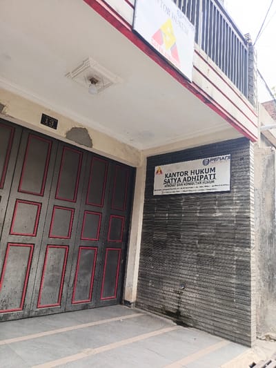 The exterior of a building features a large gray door with red trim and a plaque with the number "13" above it. A sign above the door reads "KANTOR HUKUM SATYA ADHIPATI" in Indonesian, indicating a law office. Another smaller sign with the same name and the word "PERADI" is affixed to a textured gray wall to the right of the door. A simple outdoor light is set in a square decorative fixture above the door.