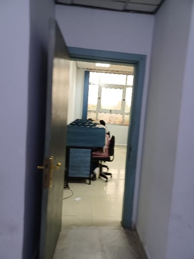 The image depicts an interior scene observed through a partially open dark-colored door with a brass-toned handle, located in Ash-Shaghour Municipality, Syria. The view is into a brightly lit office space. The floor of the entryway consists of a darker-toned threshold leading to a light-colored tiled floor within the office.

Within the office, multiple modular workstations with teal/blue partitions are visible. At least one individual is seated at a desk, facing away from the camera, and appears to be wearing dark clothing. Two office chairs are present, one dark, and another with a dark base and a reddish-brown backrest. Loose wires and scattered white papers are visible on the tiled floor.

The far wall of the office features a large multi-pane window, allowing significant natural light into the room. A light blue curtain is visible to the left of the window. The exterior scene visible through the window is bright and somewhat indistinct, suggesting daylight. The walls inside the office are white. The ceiling is equipped with visible light fixtures.