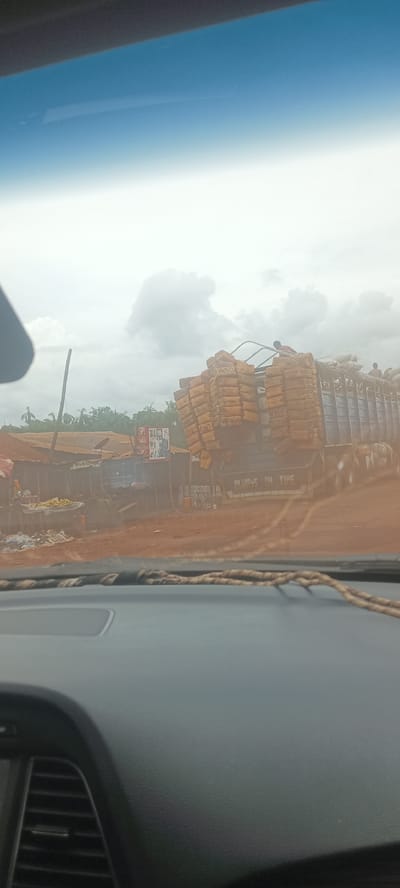 From the perspective inside a car, the image shows a truck heavily loaded with packages in Oto, Nigeria. The truck, with "Always on Time" written on its side, is packed beyond capacity. People are visible on top of the load, suggesting a lack of safety precautions. Adjacent to the road is a makeshift roadside setup with tables and goods for sale, likely a local market. The environment appears rural, with trees and foliage visible in the background, and the sky is overcast. The overall scene depicts typical conditions in a developing area, reflecting transportation and commerce.