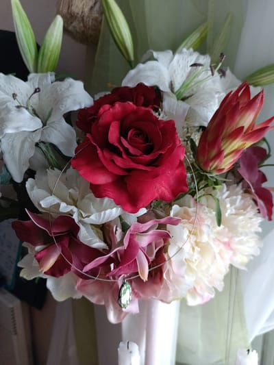 A still life photograph captures a bouquet of artificial flowers, predominantly white lilies and a deep red rose, accented by red and pinkish-purple orchids and a red protea. A delicate silver wire necklace with a green pendant is artfully draped across the bouquet, resting among the petals. The arrangement is presented in a pale pink vase, partially visible at the bottom center of the frame. The background is softly blurred, suggesting an indoor setting, with hints of green fabric and wooden textures. There are no people present in the image. The scene is static and posed, with no observable actions or interactions. The lighting appears to be natural, likely coming from a nearby window, casting soft shadows. The focus is sharp on the central elements of the bouquet and necklace. There is no information available to suggest the location in Évreux, France.