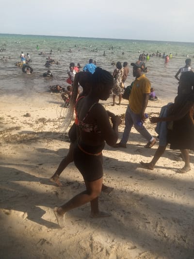 The image depicts a crowded beach scene in Shanzu ward, Kenya, under clear daylight conditions. A light brown sandy beach, marked by footprints and scattered dark organic debris, transitions into a calm, greenish-blue ocean.
In the foreground, two females with dark skin walk barefoot on the sand, moving from left to right. The leading female wears a patterned bikini top (orange, pink, black), dark shorts, and a visible beaded waist chain; her hair is styled in long braids, some featuring light-colored extensions. The second female, partially obscured behind the first, also wears a patterned bikini top and dark bottoms.
In the midground, a male subject with dark skin, wearing a yellow short-sleeved t-shirt, light blue trousers, a dark baseball cap, and sandals, walks from left to right. He carries a dark shoulder bag and holds a blue object in his left hand. Other individuals are present on the beach, including a female in a patterned dress and another in a blue top standing near the water's edge.
The ocean contains numerous individuals, estimated between fifty and seventy, primarily of African descent, engaged in swimming, wading, and floating. Many are utilizing black inner tube flotation devices. The overall scene indicates a busy public beach environment.