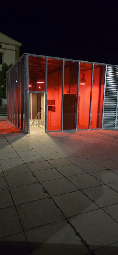 The image shows the exterior of a modern building at night. The building features a glass facade with an interior illuminated by a vibrant red light. The structure is multi-leveled, with horizontal metallic frames dividing the glass sections. Inside, one can see light fixtures, vents, and some framed items on the wall. Through the glass, a staircase is visible leading upwards. To the left of the building, a section of another structure, possibly older and lighter in color, is partially visible. The ground in front of the building is paved with large, square tiles. The scene is dark, suggesting it is nighttime, and the red lighting creates a dramatic and possibly inviting atmosphere. The location is Huesca, Spain.