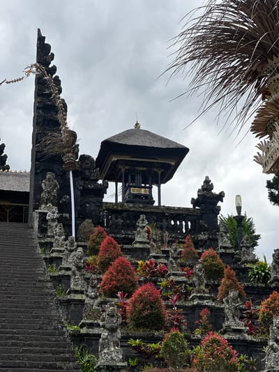 The image shows a portion of Pura Besakih temple complex in Rendang, Indonesia. The temple structure is primarily constructed of dark stone, with a thatched roof on a smaller building within the structure. Parts of the temple are adorned with red and green plants. A white cloth banner is visible on one of the structures. The cloudy sky fills the background. Some palm fronds appear on the top left. The image appears to be taken from a low angle, looking up towards the temple structure. Stone steps lead towards the temple.