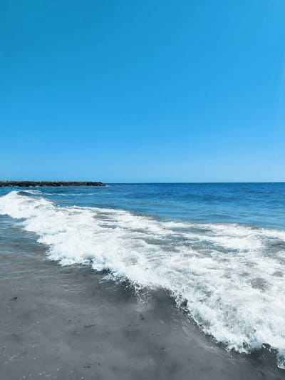 The image depicts a bright, clear day at the beach in Rota, Spain. The sky is a vibrant, cloudless blue, dominating the upper portion of the frame. The sea stretches out to the horizon, a deep blue that transitions to a lighter shade closer to the shore. A gentle wave is breaking in the foreground, its white foam creating a stark contrast against the darker, somewhat murky water and the sandy seabed, which is visible through the shallow water and contains small dark particles. In the distance, a man-made structure, possibly a jetty or a breakwater made of dark stones, extends from the left side of the image towards the right, cutting across the horizon line. There are no people or animals visible in the scene. The overall impression is one of a serene and natural coastal environment.