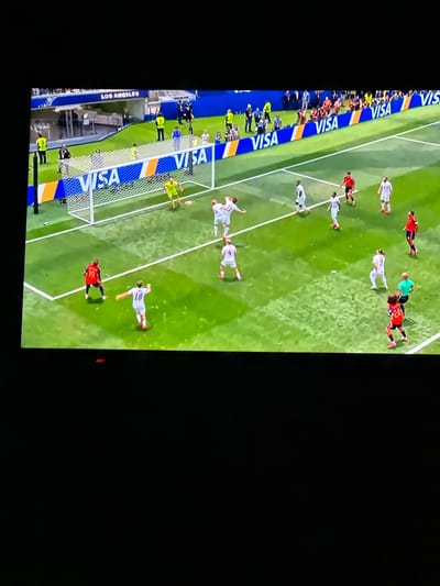 This is a medium shot of a soccer game in progress. The image appears to be captured from a television screen, as indicated by the black border around the scene. A large white net goal with "VISA" branding is prominent in the center left of the frame. Several soccer players in white and red uniforms are on the green field, some actively playing and others standing. Spectators are visible in the background behind a blue barricade, and further in the distance, banners with "LOS ANGELES" are displayed, suggesting the game might be taking place in a stadium that hosted the Olympics or another major international event. The lighting suggests it is daytime.