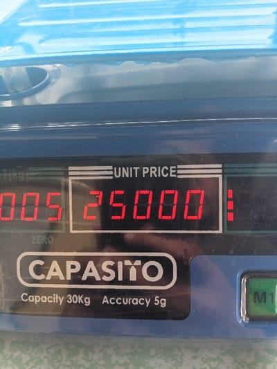 A close-up shot shows a blue digital scale. The display reads "00.5000" for weight, with "025.000" under "UNIT PRICE." The brand name "CAPASITO" is printed on the scale, with "Capacity 30kg Accuracy 5g" below. A "ZERO" indicator is visible. A green "M1" button is located in the upper right corner of the scale. The background is blurred.