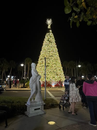 A nighttime shot shows a tall, brightly lit Christmas tree adorned with numerous lights and topped with a star. In the foreground, a white marble statue stands on a pedestal. People stroll by the tree and statue on a paved path illuminated by streetlights. The scene is framed by dark foliage on the left and additional trees and lights in the background, creating a festive and illuminated atmosphere.
