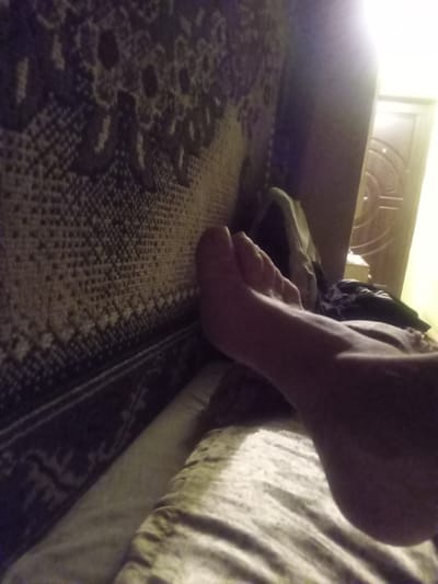 The image shows a close-up view of a person's foot resting on a textured surface, likely a bed or sofa, with a patterned fabric in the background. To the right, a door with a circular design is visible, illuminated by a bright light source. The scene appears to be indoors, possibly a bedroom or living area. There is no indication of the location being in Tiachiv, Ukraine, or any other specific city. The image is focused on the foot and the surrounding textures, with limited detail of the environment.