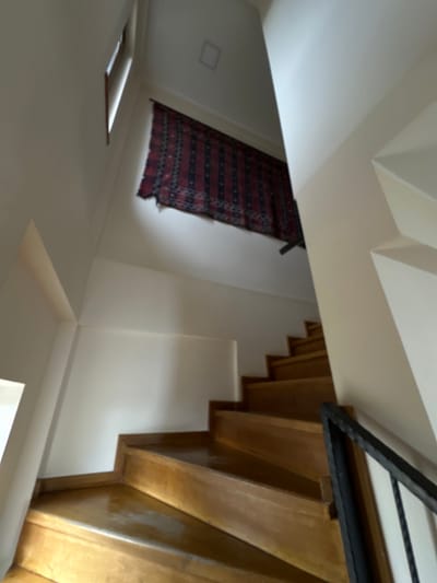 The image presents an interior view of a multi-level space, specifically a stairwell, located in Sighnaghi, Georgia. A polished wooden staircase with multiple treads and risers ascends from the lower left foreground, making a turn to the right and continuing upwards out of the frame. The walls are uniformly painted in a light, neutral color, consistent with an off-white or cream tone. High on an upper wall, positioned above the visible turn of the staircase, a rectangular textile, identified as a dark red and black patterned rug or tapestry with fringed edges, is horizontally suspended. A narrow, vertical window with a dark frame is visible on the upper left wall, admitting natural light. On the ceiling, towards the upper center, a recessed square fixture is visible. A black metal handrail with vertical balusters is partially present along the right edge of the staircase. No individuals or specific actions are depicted within the scene.