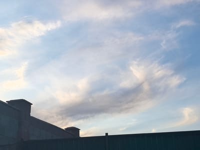 The image shows a view of the sky with clouds above a dark green fence and a dark-colored building with chimneys. The sky is a clear blue with wispy, light-colored clouds that appear to be illuminated by the sun. The clouds have a textured, feathery appearance, with some areas appearing brighter and more opaque, while others are translucent. The building in the foreground is silhouetted against the sky, with its dark color and the shapes of the chimneys providing a stark contrast. The fence is also dark and spans horizontally across the bottom of the image. The overall impression is of an outdoor scene, likely a rooftop or elevated view, with the sky as the dominant subject. The lighting suggests it might be late afternoon or early evening, possibly during a time of fair weather. The location is identified as Taldykorgan, Kazakhstan. There is no visible text in the image.