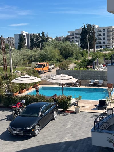 In a courtyard adjacent to a swimming pool, a black Audi sedan is parked on a paved area. To the left of the Audi, a red motorcycle is positioned. The swimming pool is surrounded by greenery and lounge chairs. Two individuals are visible near the pool, one standing and partially in the water, and another walking away from the camera. In the background, an orange truck is parked on a road, with several modern apartment buildings visible behind it. The sky is clear and blue. The scene appears to be in the city of Ulcinj - Ulqin, Montenegro.