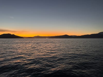 The image captures a serene sunset over the Bay of Angra dos Reis, Brazil. The sky transitions from a deep blue at the top to a vibrant orange and yellow band just above the horizon. Silhouetted against this colorful sky are a series of dark, undulating mountains that form the coastline. The water in the foreground is calm, with gentle ripples reflecting the warm hues of the sunset, creating streaks of orange and gold on the dark surface. In the distance, along the base of the mountains, a faint line of lights suggests a settlement or town. The overall impression is one of natural beauty and tranquility.