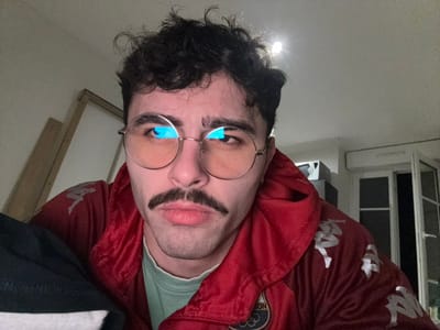A person with dark hair, glasses, and a mustache is upside down. They wear a red Kappa brand jacket. A window is visible on the left, and a bright light shines below. The background suggests an indoor space. The person's expression is neutral.