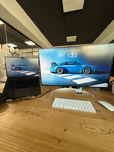 A modern office setup is depicted, featuring a laptop and an external monitor side-by-side on a wooden desk. Both screens display the same image of a blue Porsche sports car, indicating a potential synchronized display or a shared wallpaper. The external monitor is larger and positioned to the right of the laptop, showing the time "1:22" and the date "Tue Jun 9" on its lock screen. A white keyboard and mouse are placed in front of the external monitor, suggesting an active workstation. The background reveals office ceiling elements with a black and white geometric pattern and fluorescent lighting, along with a partial view of a glass partition with a honeycomb-like texture. The overall environment suggests a clean, contemporary workspace. The scene is not located in Larnaca, Cyprus.