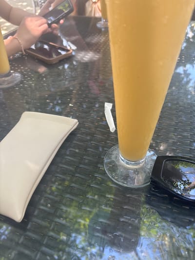 The image shows a close-up of a tall glass filled with a yellow, possibly fruity, beverage. The glass is placed on a table with a woven texture, likely a patio table, set outdoors. Sunlight reflects off the glass and the table surface, suggesting it is daytime. In the background, out-of-focus greenery and foliage indicate a natural setting, possibly a garden or tropical environment.
Visible objects include the glass of drink, a clear glass coaster, a small white paper wrapper, a pair of sunglasses with a reflection of trees and sky, and two mobile phones. One phone is held by a person's hand, which is also adorned with a bracelet. Another phone rests on the table. A light-colored object, possibly a purse or a case, is also on the table. The overall mood seems relaxed and casual, implying a leisurely activity like enjoying a drink outdoors. The light suggests it's a bright day. The mention of Samal, Philippines, as the location context points to a warm, possibly humid, tropical climate. No visible text is present in the image.