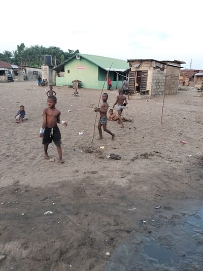 A group of children play on a sandy, littered area. Several are shirtless, and one is holding a stick. Buildings of varying construction are visible in the background, including a green building with a speaker on the roof. The sky is overcast. In the foreground, a dark patch of ground with water is visible.