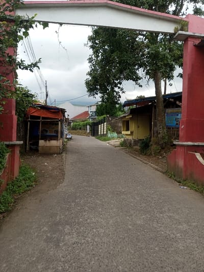 The image displays a paved road in Tagtse Tashidingkha, Bhutan, observed through a red and white archway. On the left, a small, open stall with an orange tarp roof and wooden supports is situated beside the dirt shoulder, featuring a partially legible sign that includes "Nasi Ayam". Overhead, communication lines traverse the frame. On the right, a closed yellow wooden structure stands next to a large tree and a blue rectangular sign affixed to a pole. The asphalt road, devoid of individuals or vehicles, extends into the background, flanked by buildings and fences. Distant hills or mountains are visible under an overcast sky.