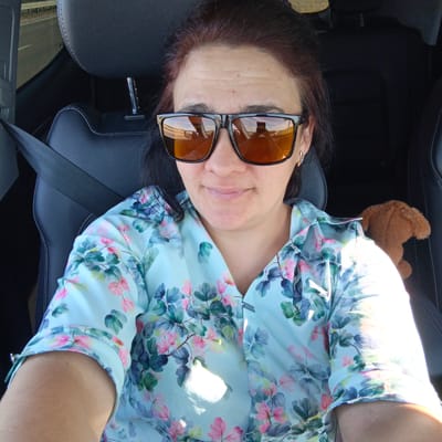 A woman with auburn hair is wearing sunglasses and a floral print shirt. She is seated in the passenger seat of a car, with a seatbelt fastened. A small brown stuffed animal is visible behind her shoulder. The interior of the car is dark leather. The background shows a road and some trees, suggesting the car is in motion. The image appears to be a selfie taken by the woman. There is no information in the image to suggest a location in Bulgakovo, Russia.