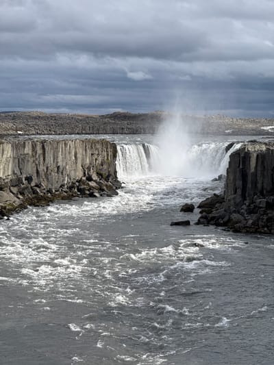 The image captures a powerful, wide waterfall in a rugged, natural landscape. A massive volume of water cascades over a dark, rocky precipice, generating a significant amount of white spray and mist that rises high into the air, partially obscuring the base of the falls.

To the right, the river flows downstream with turbulent, frothing grey water, reflecting the overcast sky above. The banks and cliffs surrounding the waterfall are composed of dark, angular rock formations. Prominent hexagonal basalt columns are visible along the left bank and extending into the middle foreground, forming stepped structures that meet the churning water.

The sky is cloudy, with streaks of grey and white clouds indicating an overcast or partly cloudy daytime. The lighting illuminates the white foam of the water and mist, creating a stark contrast with the dark rock. The scene conveys a sense of immense natural power and grandeur. No people or visible text are present in the image. The geological features, particularly the basalt columns, strongly suggest a volcanic landscape.