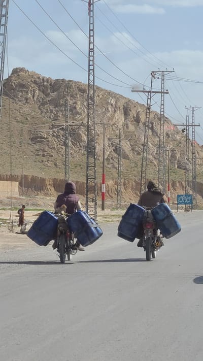 The image depicts two individuals riding motorcycles on a paved road through an arid, mountainous landscape. Each motorcycle is heavily loaded with multiple large, blue plastic barrels, suggesting the transport of liquids or other goods. The person in the foreground is dressed in maroon/purple clothing, while the one behind wears brown. A small child stands near the second motorcycle. The scene is brightly lit under a clear sky with scattered clouds, indicating daytime.

The background is dominated by rocky, barren mountains. A complex network of power lines and their supporting structures (pylons and lattice-like towers) extends across the terrain, some running alongside the road and others ascending the mountain slopes. A red and white striped barrier is visible in the mid-ground. Further in the distance, a blue sign with white text, likely in Urdu or a similar script, is partially visible. This arid and rugged environment, coupled with the reliance on motorcycles for heavy transport, is characteristic of rural or semi-urban areas in regions like Kuchlak, Pakistan. The image captures a moment of daily life and commerce, highlighting the adaptation of transportation methods to the local geography and infrastructure.