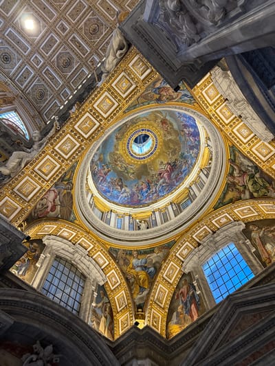 This is an upward-angled interior view of St. Peter's Basilica in Città del Vaticano, Vatican City, showcasing its grand dome and surrounding ceiling structures. The central feature is a massive circular dome, painted with a celestial scene depicting numerous robed figures, possibly angels and saints, in vibrant blue, red, and gold hues. An oculus at the dome's apex admits natural light, bordered by a blue band with gold Latin inscriptions.
Radiating from the dome are sections of barrel-vaulted ceilings and arches, heavily adorned with intricate gold ornamentation, white square panels featuring small religious motifs (such as a white dove), and additional large-scale frescoes. These murals depict various robed figures, some with books, a figure with a fish-like creature, and another with a trumpet. White marble statues of draped figures, one appearing to hold a large key, are positioned along architectural ledges. Further back, a flat or slightly curved coffered ceiling, intricately patterned with gold and white geometric designs, is visible. Several arched and rectangular windows, some displaying a clear blue sky through a grid pattern, are integrated into the architecture. Artificial spotlights illuminate various sections of the ornate ceiling. The dominant colors are rich gold, white, and the vivid blues, reds, and greens of the frescoes, all contributing to an opulent and grand atmosphere.