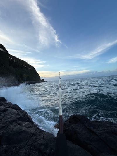The image captures a coastal rock fishing scene in Hitiaʻa ʻo te Rā, France. In the immediate foreground, dark, rugged volcanic rock formations are visible. A light-colored fishing rod with a dark handle extends diagonally from the lower center-right, with its line cast into the turbulent, deep blue-green ocean water. To the left, large waves forcefully crash against the rocks, generating prominent white spray and foam. Further left, a steep hillside densely covered with dark green vegetation rises sharply from the water's edge; a narrow, dark strip at its base suggests a coastal road or path. The ocean dominates the midground and background to the right, extending to a horizon where faint landmasses or low-lying clouds are discernible. The sky is partly cloudy, transitioning from a lighter blue near the horizon to a deeper blue overhead, with several elongated white clouds present. The lighting conditions suggest either early morning or late afternoon. No individuals are visible in the scene.