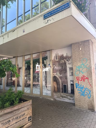 The image shows an outdoor scene with a modern building featuring a large glass facade. Above the glass, a concrete structure is covered with graffiti. The graffiti includes abstract shapes in blue and red, as well as some illegible tags. Below the concrete structure, a mural depicting a brick building facade with arched doorways is painted on the wall, extending across the glass. The glass facade reflects the surroundings, including trees and the sky.

To the left of the building, a blue street sign reads "Meerminneplein". In front of the building, a wooden planter box with plants is visible, and a brick-paved sidewalk runs alongside it. The planter box has text stenciled on its side. Part of the text is legible and reads "Tijdelijk stukje groen!" and "Zwolle". Below this, a URL is partially visible, reading "www.j-p-l-kunstenaars-collectief.nl/groen".

The overall impression is an urban setting that combines modern architecture with artistic elements like graffiti and murals. The image appears to have been taken during the daytime, under clear skies, judging by the reflections in the glass and the lighting. There are no people visible in the scene.