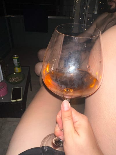 The media file shows a close-up, angled shot of a hand holding a large wine glass partially filled with amber-colored liquid. The glass is tilted, with some condensation visible on its exterior. The background is blurred, but suggests an indoor setting with dim lighting. To the left, there are hints of decorative lights. In the lower part of the image, a table is visible with a few items on it, including a can, a small bowl with some contents, and a smartphone. A small glass with text is also visible on the base of the wine glass stem. The overall lighting suggests it is likely evening or night. The setting appears to be a casual gathering or personal space. The visible text on the glass stem reads "Vino by Riedel".