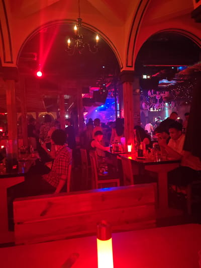 The image depicts the interior of a nightclub or bar located in Xuanwu District, China. The scene is dominated by artificial lighting, primarily deep red in the foreground and shifting to blue and purple in the background.
Architectural elements include multiple arched doorways or passageways with dark trim, suggesting a structured interior design. In the upper left, a red spotlight beam projects across the archway, and a dark-colored chandelier with four candle-like lights is suspended from the ceiling. Further back, another larger chandelier, possibly white or light-colored, hangs above a different section. Wooden pillars and beams are visible throughout the space, defining separate areas.
Approximately two dozen individuals, appearing to be adults, are present in various parts of the venue. In the mid-foreground, a person with dark, curly hair, wearing a red plaid shirt, is seated at a table, facing away from the viewer. Another individual in a patterned top is standing near a bar on the left. On the right side of the midground, a group of three to four men are seated at a table, with one man in a light-colored shirt looking at a smartphone. Other individuals are standing and conversing in groups throughout the central and background areas, some holding drinks or phones.
Various objects are distributed across the scene. Dark-colored tables are equipped with glasses and bottles. On the left, a large clear pitcher filled with a dark liquid sits on a bar counter. In the immediate foreground, a dark red table surface is visible, upon which rests a cylindrical table lamp emitting a warm yellow-orange light. A green and white exit sign with an arrow pointing right is mounted high on a wall in the midground. The background features more extensive lighting installations, including what appears to be decorative string lights or small spot lights. The overall environment suggests a social and active nightlife setting.