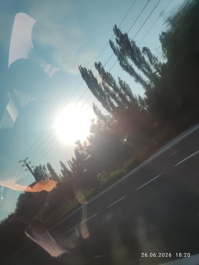 The image captures a dynamic scene from a moving vehicle, likely on a road trip, during a bright, sunny day. The perspective is from inside the car, looking out at the landscape blurring past.

The primary subjects include a road with white lane markings, suggesting a highway or main route. Trees and vegetation line the side of the road, appearing as streaks of dark green due to motion blur. Overhead, power lines crisscross the sky, supported by a utility pole visible on the left. The sky is a clear, bright blue, with the sun prominently positioned, casting a strong glare and creating lens flare effects. There's a reflection on the windshield, showing parts of the car's interior, including what appears to be the driver or passenger.

The activity depicted is travel, evidenced by the motion blur and the road ahead. The time of day is late afternoon, as indicated by the timestamp "18:20" and the bright, low angle of the sun. The weather is sunny and clear.

Notable details include the strong lens flare from the sun, the motion blur of the surrounding trees and landscape, and the reflection on the windshield. The timestamp "26.06.2026 18:20" provides specific temporal information. While no specific emotions are directly conveyed, the bright sunlight and open road can evoke feelings of freedom, journey, or anticipation. The location context provided is Yanaryk, Uzbekistan.