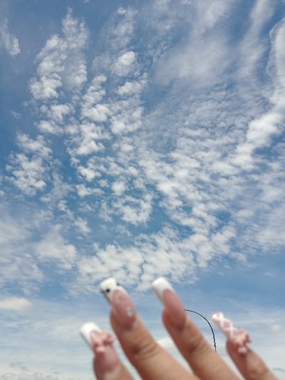 A human hand is prominently featured in the blurred foreground of an upward-angled outdoor photograph, set against a vast blue sky filled with numerous scattered white clouds. The image was captured in Germiston, South Africa. The hand displays long, manicured nails with white French tips. Specific adornments include a light pink 3D bow on the thumb and index finger nails, a small clear gemstone and a white 3D embellishment on the middle finger nail, and a light pink 3D criss-cross pattern and bow on the pinky finger nail. A slender, dark, curved object is faintly visible in the blurred mid-background between the ring and pinky fingers. The sky exhibits varying patterns of cumulus or stratocumulus clouds under bright daylight conditions.