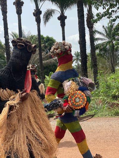 In Agba Umana, Nigeria, two masquerades stand outdoors on a sunny day with palm trees visible. One masquerade wears a black net-like mask adorned with small, round objects, possibly shells or nuts. It holds a red, elongated object. The other masquerade is more colorful, featuring a striped, knitted body, a circular headdress with a patterned design, and various hanging materials.