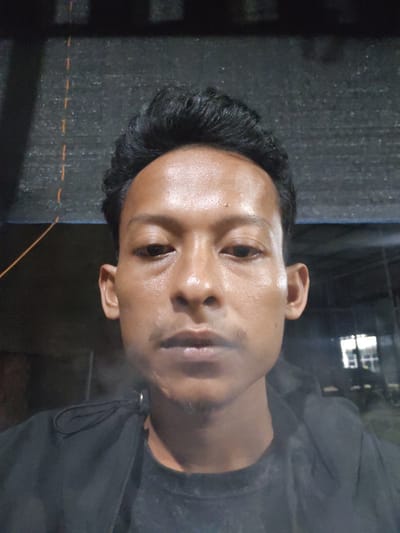 A close-up, head-and-shoulders selfie of a young man is captured in a dimly lit setting. He is wearing a black hooded jacket over a dark t-shirt. His dark hair is styled upwards. His face is illuminated, highlighting his skin which appears slightly sweaty or oily, and his features including his eyes, nose, and lips. He has a faint mustache and stubble on his chin. The background is dark and out of focus, with some structural elements and what appears to be a grate or mesh pattern visible above him. To the right, there are some illuminated rectangular shapes, possibly windows or lights, suggesting an indoor or covered outdoor space. The lighting suggests it could be nighttime or a dark indoor environment. No visible text is present in the image. The overall impression is a casual, informal self-portrait.