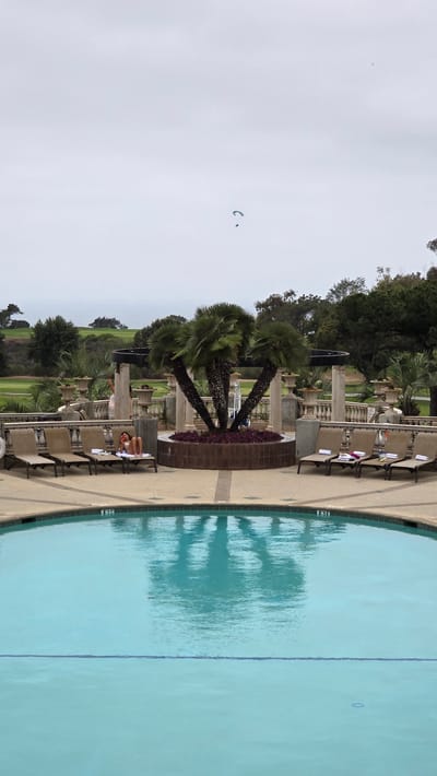 A person is reclining on a lounge chair by a swimming pool, appearing to eat a piece of fruit. The pool area is paved and features several other empty lounge chairs, each with towels. A circular planter with palm trees stands prominently in the background, with an ornate stone balustrade and columns surrounding it. Beyond the pool area, a golf course is visible, with rolling green hills and trees under a cloudy sky. In the distance, a paraglider is visible in the sky. The image appears to be taken in La Jolla, San Diego, United States of America.