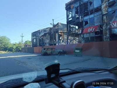 The image shows the aftermath of a fire in an urban environment. Two multi-story buildings on the right side of the frame are heavily damaged, with blackened facades and shattered windows. A red sign with white lettering, partially obscured by soot, is visible on the front of one building. The building on the left is also extensively damaged, with a partially collapsed roof and exposed wooden beams.

In the foreground, a burnt-out SUV is parked on a paved surface. Next to it, a green dumpster with "CLEAR CITY" written on its side stands near the damaged buildings. The ground is covered in debris and dust. The scene suggests destruction and abandonment.

The sky is clear and blue, indicating daytime, likely late afternoon given the timestamp at the bottom right corner: "22.06.2026 17:33". The presence of power lines and trees in the background suggests an urban or semi-urban setting. The overall mood of the image is somber, conveying the devastating impact of the event. The location context provided is Lymanka, Ukraine.