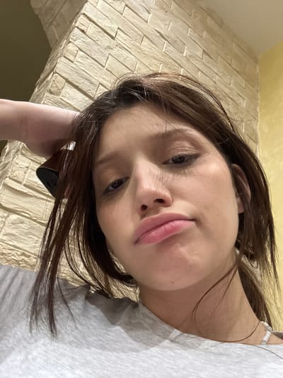 A young woman with dark hair, wearing a grey t-shirt, poses for a selfie. Her left arm is raised and resting on her head, holding a dark-colored object. She has a pouty expression with her lips slightly pursed. The background shows a textured wall that appears to be made of light-colored bricks, and a yellow-painted wall to the right. There are no discernible landmarks that indicate the location is Surgut, Russia.