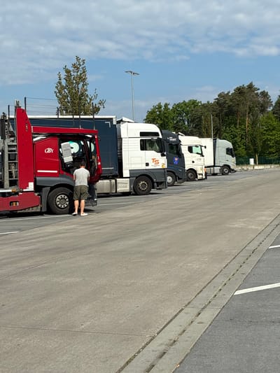 The image shows a row of parked semi-trailer trucks in Baar-Ebenhausen, Germany. A person is standing next to the front truck, a red vehicle with "GBY" printed on the side. The trucks are parked on a concrete surface with painted white parking lines. The background includes trees and a blue sky with clouds.