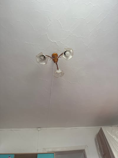 The image shows an interior view of a ceiling with a chandelier. The ceiling is painted white and has a textured, cracked surface. A three-armed chandelier with ornate golden metalwork and white floral-shaped lampshades is visible in the upper center of the frame. The lampshades contain light bulbs. The bottom portion of the image shows a white wall, a doorway, and a part of a cabinet with teal accents. The scene is likely a residential interior in Yabogan, Russia, as specified. There are no people or animals present in the image.