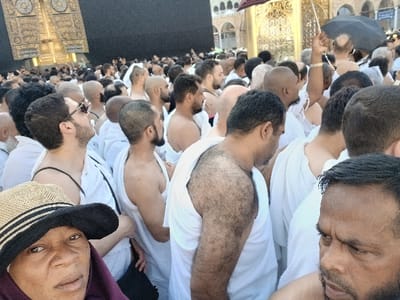 The image depicts a crowded scene within the Al-Masjid al-Haram in Makkah Al Mukarramah, Saudi Arabia. In the upper left background, a portion of the Kaaba is visible, characterized by its black covering and ornate golden door. A dense congregation of individuals, predominantly men, occupies the foreground and mid-ground. Most men are dressed in white Ihram garments, with some having one shoulder uncovered. Visible features among the men include various beards, shaved heads, and closely cropped hair. In the lower left foreground, a woman with dark skin, wearing a straw hat with a black band and a dark head covering, is looking directly towards the viewer; she has a nose piercing. To her right, a man with a beard and dark hair is partially visible, looking downwards. In the middle ground, a man is seen holding a dark umbrella. The crowd appears to be standing or moving slowly within the sacred precinct.
