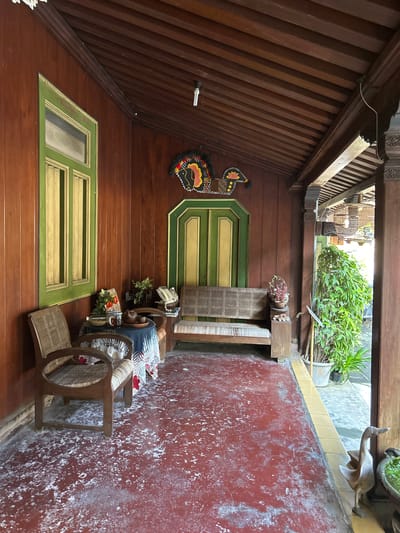The image depicts an outdoor courtyard or veranda of a traditional Indonesian home, likely in Banguntapan, Indonesia. The scene is characterized by its rustic and antique aesthetic, with abundant use of dark, polished wood for walls, ceilings, and architectural details. A slatted wooden ceiling angles upwards, revealing more wooden beams and decorative elements.

Dominating the left side of the frame are two prominently placed wooden window frames, painted a vibrant green with yellow trim. Above the upper window, a colorful, intricately designed applique of a bird or butterfly is affixed to the wooden wall.

The right side of the image features a weathered, reddish-brown wall with white splotchy discoloration, suggesting age or perhaps a unique decorative finish. Adjacent to this wall is a wooden chair with a woven rattan seat and back, occupied by a figure. The figure is partially visible, wearing a dark blue garment and a patterned fabric draped over their lap. Nearby, a small wooden table holds what appears to be books or magazines and a potted plant. Further into the courtyard, a longer, bench-like seat, also constructed from woven rattan and wood, is visible.

The background reveals a glimpse of lush greenery from potted plants, indicating an outdoor setting. The lighting suggests it is daytime, possibly late morning or early afternoon, with natural light illuminating the space. The overall mood is one of tranquil, traditional living, with a sense of history and cultural heritage. There is no visible text in the image.