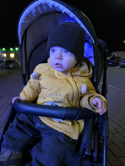 A young child, approximately a toddler, is depicted seated in a black baby stroller at night in the city of Voskresensk, Russia. The child wears a dark brown knit beanie, a yellow padded jacket featuring a light-colored graphic design and two beige circular embellishments on the shoulders, and dark blue jeans. Their blue eyes are open and directed towards the right side of the frame, with their mouth slightly ajar. The scene is illuminated by prominent blue ambient light, which casts a cool, blue hue across the child's skin and visible stroller components. The stroller features a black frame and a padded handlebar. In the blurred background, a building with illuminated windows and a string of green decorative lights is visible to the left, while multiple parked vehicles are discernible in a dark street or parking area. The ground is paved with light-colored bricks.