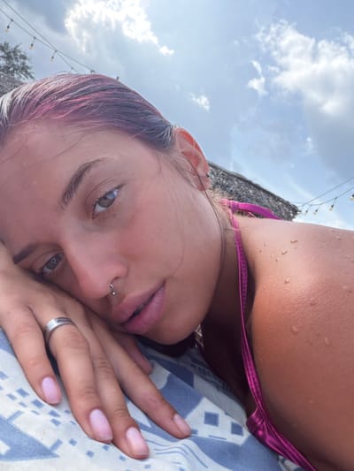 A woman with pink-streaked hair lies on a patterned fabric, her head resting on her hand. She wears a vibrant pink bikini top. Water droplets are visible on her skin. Her left hand, adorned with a silver ring, is positioned near her face, showcasing her light pink fingernails. Her right hand is not visible. The background features a glimpse of a thatched roof, string lights, and a cloudy sky, suggesting an outdoor, possibly tropical, setting. The image captures a close-up perspective of the woman's face, with her eyes open and looking towards the viewer. The scene is set on a sunny day.