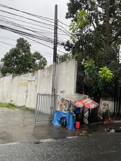 The scene takes place in Cimahi, Indonesia at the coordinates -6.886 latitude and 107.554 longitude. A street vendor is standing behind a food cart, which is covered by a blue tarp and a red awning. There is a metal gate to the left of the vendor and a tall concrete wall behind them. There are multiple power lines crisscrossing overhead and several trees in the background. The ground is wet from recent rainfall.