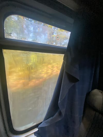 This image captures a view from inside a vehicle, looking out of two windows. The windows appear to be slightly fogged or wet, with streaks and condensation visible, obscuring the details of the exterior. The left window shows a blurred, impressionistic scene of what might be trees or foliage with patches of bright light, suggesting either dawn or dusk, or passing through a brightly lit area. A small circular red and white sticker with a symbol is visible on this window. The right window is largely covered by a dark blue curtain, which is partially drawn back, revealing more of the fogged glass. The curtain is draped and appears to be made of a fabric with a subtle checkered pattern. The interior of the vehicle is dark, with the texture of a seat visible in the lower right corner, covered by the curtain. The overall impression is one of travel, with a sense of being enclosed within the vehicle while observing the outside world through a diffused lens. The location in Bojišta, Montenegro, is not directly evident from the visual cues in the image itself, but this context is provided externally.
